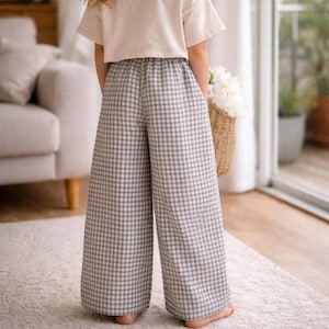 Pode incluir: Uma pessoa vestindo cal&ccedil;as largas xadrez cinza e branco e uma blusa creme. A pessoa est&aacute; segurando uma cesta de vime cheia de flores brancas. As cal&ccedil;as t&ecirc;m cintura el&aacute;stica.