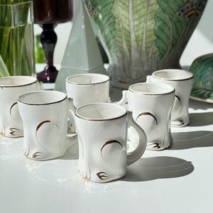 Puede incluir: Seis tazas de cerámica blanca con ribetes y detalles dorados. Cada taza tiene una forma única con un asa y detalles dorados decorativos. Las tazas están dispuestas sobre una superficie blanca, sugiriendo un juego para servir bebidas.