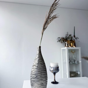 Può includere: Un vaso argentato e strutturato con una forma alta e sottile. Il vaso contiene uno stelo di pianta marrone secco. Una candela decorativa in un bicchiere e un mobile sono sullo sfondo. Gli oggetti sono su una superficie bianca.