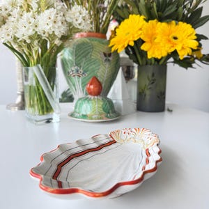 Pode incluir: Um prato de cerâmica branco em forma de concha, com listras vermelhas e pretas e uma borda vermelha. O prato está sobre uma superfície branca, com vasos de flores ao fundo.