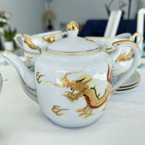 Op de afbeelding: Witte porseleinen theeset met gouden rand en een drakenmotief. De set bevat een theepot, kopjes en schotels. De draak is afgebeeld in goud, rood en geel. De theepot heeft een gouden knop op het deksel.