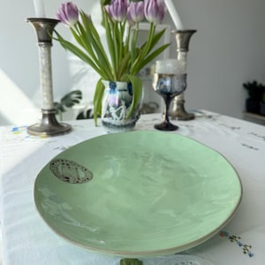 Pode incluir: Um prato de servir oval verde claro com um emblema decorativo. O prato está sobre uma toalha de mesa branca com bordados florais. Ao fundo, há tulipas roxas num vaso, castiçais e uma taça de vinho.