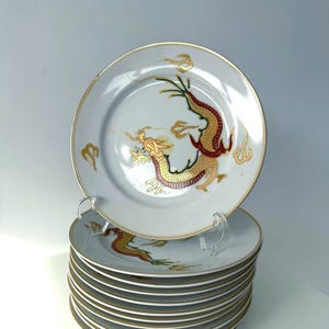 Könnte beinhalten: Ein Stapel weißer Keramikteller mit goldenem Rand und einem Drachendesign. Der Drache ist in Gold, Rot und Grün dargestellt. Die Teller sind auf einem transparenten Kunststoffständer gestapelt.