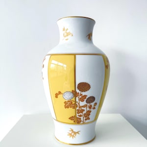 Pode incluir: Um vaso de porcelana branca com painéis verticais amarelos e motivos florais em tom dourado, incluindo crisântemos e detalhes de bambu. O vaso tem uma borda e base alargadas, com linhas douradas finas que delineiam os painéis e elementos.