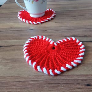 May include: Red heart-shaped crochet coasters with white trim. One coaster has a white teacup on top. The coasters are on a wooden surface. The coasters are handmade and have a decorative design.