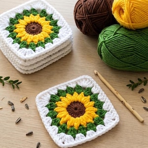 May include: A stack of three square crochet coasters and one individual coaster featuring a sunflower pattern with a brown center, yellow petals, green leaves, and a white border, accompanied by three balls of yarn and a wooden crochet hook.