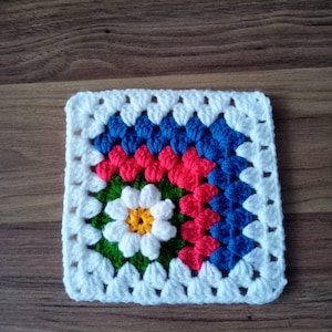 May include: A square, crocheted granny square in white, blue, red, green, and yellow. The center features a white flower with a yellow center, surrounded by green. The square is bordered in white.