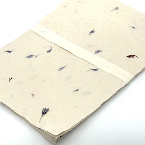 Könnte beinhalten: Ein Stapel handgefertigtes Papier mit cremefarbenem Grund, verziert mit kleinen blauen und braunen floralen Einschlüssen. Ein cremefarbenes Band umwickelt den Stapel. Das Papier hat eine strukturierte Oberfläche, was auf ein natürliches, umweltfreundliches Produkt hindeutet.