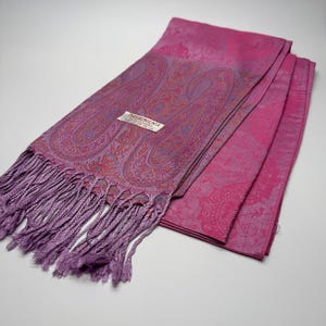 Könnte beinhalten: Ein gefalteter, magentafarbener Schal mit Paisley-Muster und Fransen. Der Schal hat ein Etikett mit der Aufschrift "Pashmina". Der Schal ist aus weichem Material gefertigt und wird wahrscheinlich um den Hals getragen.