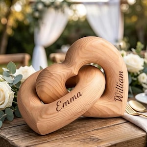 May include: Two wooden heart-shaped sculptures interlocked, with the names "Emma" and "William" engraved on them. The wood has a natural grain and a light brown color. The background includes flowers and a blurred outdoor setting.