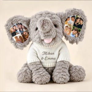 Puede incluir: Un juguete de peluche de elefante gris que lleva un suéter de punto blanco con el texto Mikan & Emma impreso en el frente. El elefante tiene orejas grandes con varias fotografías de parejas impresas y una pequeña lengua rosa visible.