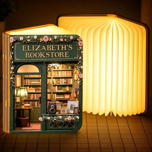 Może przedstawiać: Lampa w kształcie książki z miniaturową fasadą księgarni w kolorze ciemnej zieleni z napisem ELIZABETH'S BOOKSTORE, ozdobiona różowymi girlandami kwiatowymi. Wnętrze ukazuje półki z książkami, zieloną lampkę i ramki, emitując ciepłe światło.