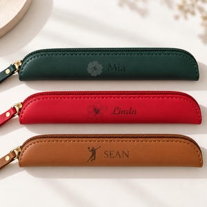 May include: Three slim, rectangular pencil cases with zippers. The top case is dark green with a flower icon and the name Mia. The middle case is red with a flower icon and the name Linda. The bottom case is brown with a volleyball player icon and the name SEAN.