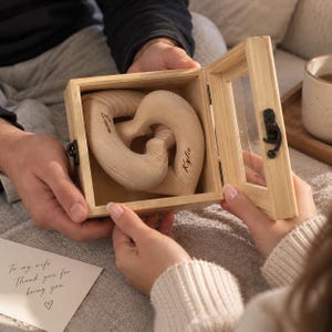 Könnte beinhalten: Eine helle Holzschatulle mit aufklappbarem Glasdeckel, die zwei ineinandergreifende, herzförmige Holzteile enthält. Jedes Herz ist mit einem Namen graviert, Ewan und Kylie, und die Schatulle hat einen kleinen Metallverschluss.