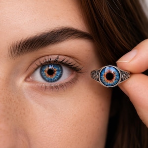 Op de afbeelding: Een zilveren ring met een ronde cabochon met een realistisch blauw en oranje irisontwerp. De ringband vertoont ingewikkeld metaalwerk met gedetailleerde krulpatronen aan de zijkanten, passend bij de kleuren van het menselijk oog.