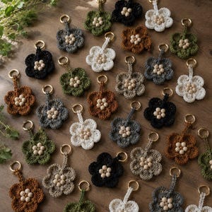 May include: A collection of crocheted flower keychains in various colors including black, white, brown, gray, and green. Each flower features a central cluster of small, light-colored wooden beads and is attached to a gold-toned metal keyring.