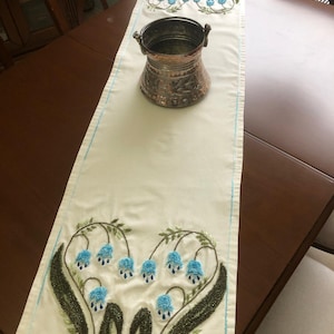 Op de afbeelding: Crèmekleurige tafelloper met blauwe bloemborduurwerk en een koperkleurige metalen container. De loper heeft een blauwe rand en ligt op een bruine houten tafel. Het borduurwerk toont blauwe klokvormige bloemen.