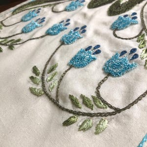 May include: Close-up of a white fabric with detailed embroidery. The design features light blue, textured flowers with dark blue accents, connected by brown stems and surrounded by green leaves. The embroidery is intricate and detailed.