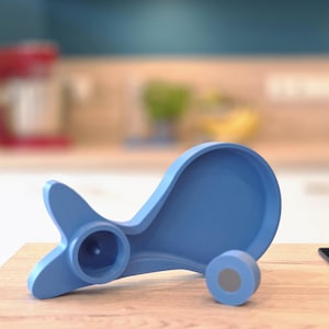 Könnte beinhalten: Ein blaues Küchenutensil mit einem einzigartigen Design, das einen kreisförmigen Sockel und eine geschwungene, flügelförmige Form aufweist. Das Utensil besteht aus einem soliden, matten Material und liegt auf einer Holzoberfläche. Der Hintergrund ist eine verschwommene Küchenszene.