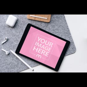 Op de afbeelding: Een digitale tablet met een roze scherm met de tekst "YOUR IMAGE HERE" en een stylus. De tablet ligt op een grijze viltachtige ondergrond, met witte draadloze oordopjes en een oplaadcase in de buurt.