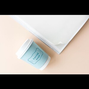 Puede incluir: Una taza de café de papel azul claro con el texto "COFFEE SHOP" en un diseño rectangular. La taza está sobre una superficie rosa claro junto a una pila de papel blanco.