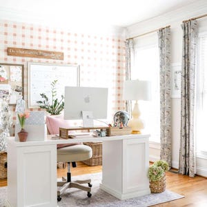 Op de afbeelding: Een lichte thuiskantoor met een witte bureau, een computer en een roze en witte geruite muur. De kamer heeft een vloerkleed, gordijnen en een lamp. Een bord aan de muur luidt: "Doe wat ertoe doet. Vergeet de rest."