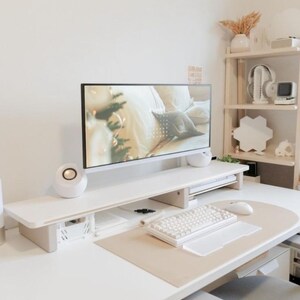 May include: A contemporary home office with a white desk, monitor, and keyboard. The desk includes a monitor stand, speakers, and a mouse. A neutral desk mat and a white keyboard are also present. A shelf with decorative items is in the background.