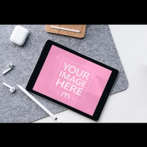 以下が含まれることがあります： 「YOUR IMAGE HERE」のテキストが表示されたピンク色の画面のデジタルタブレットとスタイラス。 タブレットは灰色のフェルトの表面にあり、白いワイヤレスイヤホンと充電ケースが近くにあります。