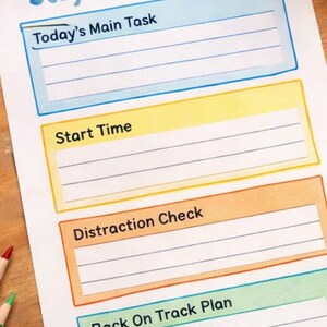 Könnte beinhalten: Ein gedrucktes "Stay On Task Chart" mit Abschnitten für "Today's Main Task", "Start Time", "Distraction Check" und "Back On Track Plan". Farbstifte und Bausteine umgeben die Tabelle. Eine blaue Uhr ist ebenfalls vorhanden.