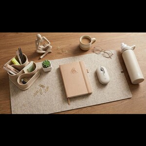 Könnte beinhalten: Ein beiges Schreibtisch-Setup mit einem Notizbuch mit Fuchslogo, einer kabellosen Maus, einer Wasserflasche, einer Kaffeetasse und einem Stifthalter. Die Artikel sind auf einer grauen Filzmatte angeordnet.