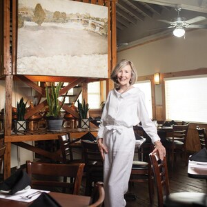 Op de afbeelding: Interieur van een restaurant met een vrouw in witte kleding. Een groot schilderij van een brug hangt aan een houten constructie. Tafels en stoelen zijn gedekt voor het diner. Rustieke sfeer.