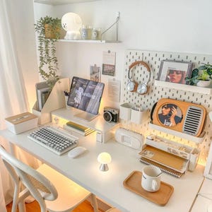 May include: A well-lit, organized workspace with a white desk, chair, and shelves. The desk features a keyboard, mouse, tablet, and a small lamp. Shelves hold plants, decor, and a radio. The word "Recording" is visible on the tablet screen.