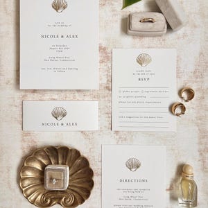 May include: A collection of wedding stationery featuring a seashell motif. Includes invitation, RSVP, and direction cards, along with a ring box, rings, and a decorative dish. The text reads "Nicole & Alex".