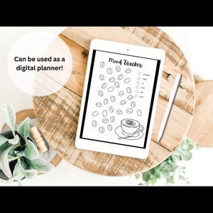 May include: A digital mood tracker displayed on a tablet, featuring coffee bean illustrations and a coffee cup. The text "Mood Tracker" is at the top. The tablet is on a wooden surface with greenery.