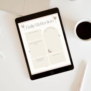 Könnte beinhalten: Ein schwarzes Tablet mit einer Vorlage für "Tägliche Reflexion" mit Abschnitten für tägliche Dankbarkeit, Absichten und Notizen. Das Tablet liegt auf einer weißen Oberfläche, neben einem Laptop, einem Stift und einer Tasse Kaffee.