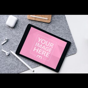 Könnte beinhalten: Ein digitales Tablet mit einem rosa Bildschirm, auf dem der Text "YOUR IMAGE HERE" und ein Stift angezeigt werden. Das Tablet liegt auf einer grauen Filzoberfläche, mit weißen kabellosen Ohrhörern und einem Ladecase in der Nähe.