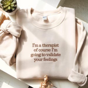 Könnte beinhalten: Beiges Sweatshirt mit dem Text "I'm a therapist of course I'm going to validate your feelings" in braunen Buchstaben. Die Ärmel sind hochgekrempelt. Im Hintergrund ist eine kleine Sukkulentenpflanze zu sehen. Das Sweatshirt ist gefaltet.