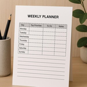 Könnte beinhalten: Ein weißer Wochenplaner mit der Aufschrift "WEEKLY PLANNER" oben. Der Planer hat Abschnitte für jeden Wochentag mit Spalten für Top-Prioritäten, To-Do-Listen und Notizen. Ein Bleistift und eine kleine Pflanze sind im Hintergrund zu sehen.