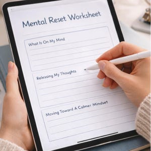 Puede incluir: Una persona usa un lápiz óptico para escribir en una tableta que muestra una "Mental Reset Worksheet". La hoja de trabajo solicita al usuario que considere sus pensamientos y avance hacia una mentalidad más tranquila. La tableta descansa sobre un libro azul.