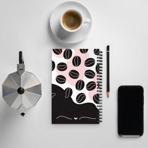 Könnte beinhalten: Ein Notizbuch mit Kaffeebohnenmuster, eine Espressotasse auf einer Untertasse, ein schwarzer Stift, ein Smartphone und ein silberner Espressokocher stehen auf einer weißen Oberfläche.