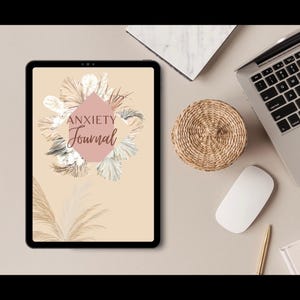 Könnte beinhalten: Tablet mit einem 'Angst-Tagebuch' mit floralem Design. Das Tagebuchcover zeigt die Worte 'Anxiety Journal' in Schriftform, umgeben von beige- und rosa-farbenen floralen Elementen. Ein Laptop, eine Maus und ein Weidenkorb sind ebenfalls zu sehen.