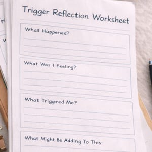 Op de afbeelding: Een stapel wit papier met de titel "Trigger Reflection Worksheet" en prompts voor zelfreflectie. Twee paperclips en een houten schaal staan op de achtergrond.