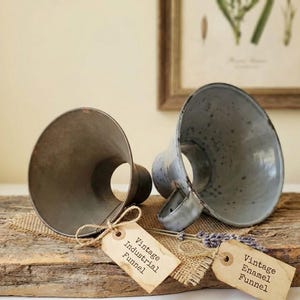 May include: Two vintage metal funnels, one industrial and one enamel, rest on a rustic wooden surface. The industrial funnel is a dark gray, while the enamel funnel is a speckled gray-blue. Each has a tag that reads "Vintage Industrial Funnel" and "Vintage Enamel Funnel".