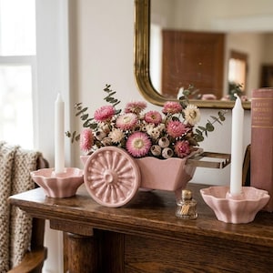 May include: A pink ceramic wheelbarrow planter filled with dried pink and white flowers. Two white candles sit in matching pink candle holders. A small glass bottle of matches is also present.