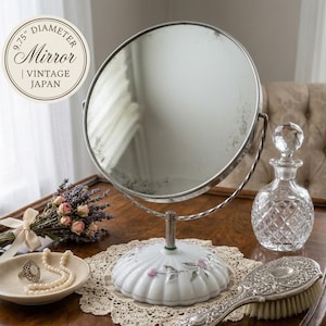 May include: A vintage vanity mirror with a 9.75" diameter, round, silver-toned frame. The white porcelain base is decorated with pink floral accents. A small dish with jewelry, a perfume bottle, and a brush are also on the table.