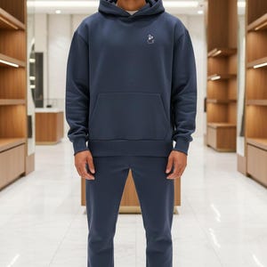 May include: Navy blue hoodie and matching tracksuit bottoms. The hoodie features a front pocket and a small embroidered design on the chest. The model is wearing brown and beige slippers with a face design. The outfit is displayed in a retail setting.