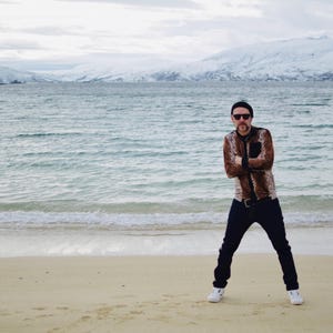 May include: A person wearing a brown and black patterned shirt, black jeans, and white trainers stands on a sandy beach. The person is wearing sunglasses and a black beanie. The background features a body of water and snow-covered mountains.