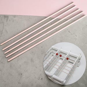 Peut inclure: Un ensemble d'outils d'artisanat comprenant cinq chevilles en bois rose clair et un métier à tisser blanc avec des fils roses et des perles rouges. Le fond est un mélange de gris et de rose.