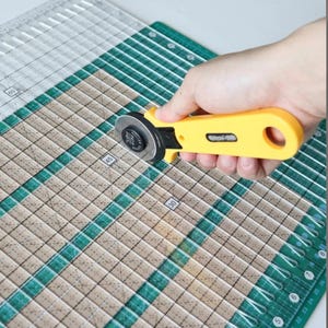May include: A yellow rotary cutter with a silver blade is positioned above a green and tan cutting mat. The mat features grid lines and measurements in centimetres. The cutter is used for fabric and paper crafts.