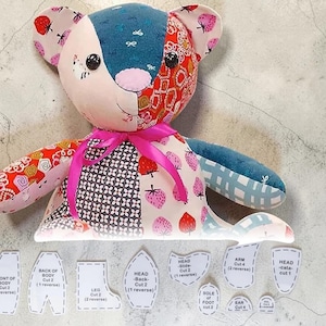 Può includere: Un orsacchiotto patchwork realizzato con vari tessuti a fantasia, tra cui fragole, forme geometriche e colori a tinta unita, con occhi a bottone neri, un naso rosa e un nastro rosa acceso legato al collo, accompagnato da cartamodelli in carta.